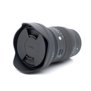 Sigma 16-28mm f2.8 Sony-1 Sigma 16-28mm f/2.8 DG DN C Sony E -objektiivi (käytetty, kunto K5, 24 kk takuu, sis. alv 24%), jossa on vastavalosuoja ja suojus, makaa kyljellään valkoista taustaa vasten.