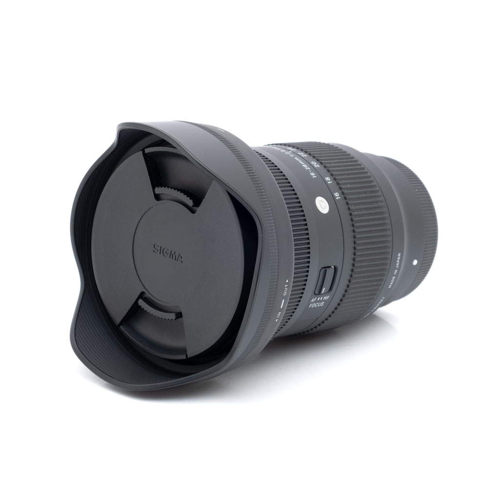 Sigma 16-28mm f2.8 Sony-1 Sigma 16-28mm f/2.8 DG DN C Sony E -objektiivi (käytetty, kunto K5, 24 kk takuu, sis. alv 24%), jossa on vastavalosuoja ja suojus, makaa kyljellään valkoista taustaa vasten.