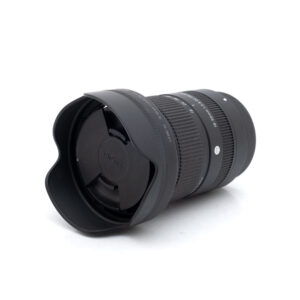 Sigma 18-50mm f/2.8 DC DN C Fuji-objektiivi (Kunto K4.5, sis.ALV24%) - Käytetty, musta, kuvassa vastavalosuoja ja etusuljin kiinnitettynä, kyljellään valkoista taustaa vasten.
