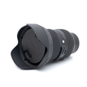Sigma 24-70mm f2.8 Sony-5269-1 Sigma Art 24-70mm f/2.8 DG DN Sony - Käytetty musta kameraobjektiivi, jossa on terälehden muotoinen vastavalosuoja ja objektiivin suojus valkoisella pohjalla, ja jossa on näkyvät kytkimet ja tuotemerkintä piipussa.