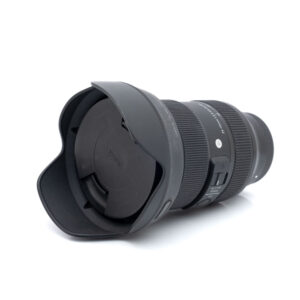 Sigma 24-70mm f2.8 Sony-5269-1 Sigma Art 24-70mm f/2.8 DG DN Sony - Käytetty musta kameraobjektiivi, jossa on terälehden muotoinen vastavalosuoja ja objektiivin suojus valkoisella pohjalla, ja jossa on näkyvät kytkimet ja tuotemerkintä piipussa.