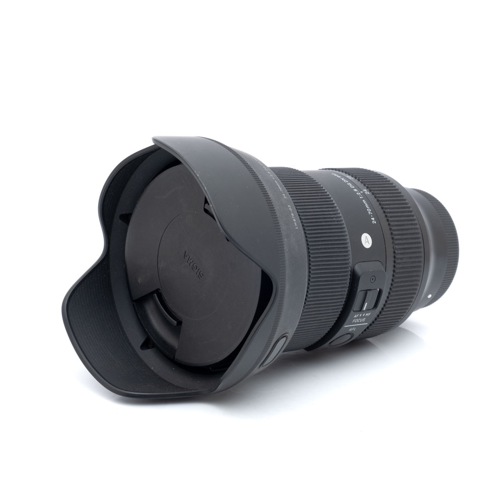 Sigma 24-70mm f2.8 Sony-5269-1 Sigma Art 24-70mm f/2.8 DG DN Sony - Käytetty musta kameraobjektiivi, jossa on terälehden muotoinen vastavalosuoja ja objektiivin suojus valkoisella pohjalla, ja jossa on näkyvät kytkimet ja tuotemerkintä piipussa.