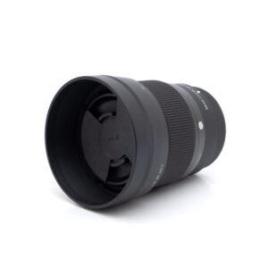 Sigma 56mm f/1.4 C DC DN Fuji (Kunto K5, sis.ALV24%) - Käytetty objektiivi, väriltään musta, jossa on vastavalosuoja ja etusuljin kiinnitettynä ja joka on kuvattuna vinossa valkoisella taustalla.