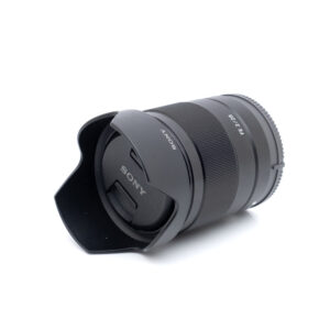 Sony FE 28mm f/2 - Käytetty objektiivi, johon on kiinnitetty vastavalosuoja ja suojus, makaa kyljellään valkoista taustaa vasten, ja siinä on tyylikäs musta muotoilu.