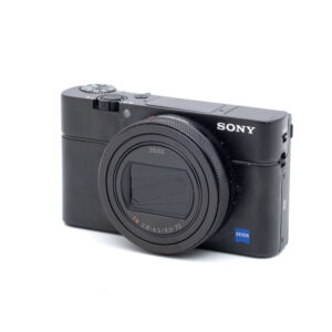 Sony RX100 VII-1 Musta Sony RX100 VII (sis.ALV24%) - Käytetty digitaalikamera, jossa on Zeiss-objektiivi, on suunnattu hieman vasemmalle valkoisella taustalla. Objektiivin suojus on suljettu ja etupuolella näkyy SONYn tuotemerkki.