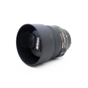 Nikon 50mm f18G-3-2 Nikon AF-S Nikkor 50mm f/1.8 G - Käytetty objektiivi, johon on kiinnitetty vastavalosuoja ja jossa on Nikonin logo etusuljessa ja kultaiset sivukirjaimet tarkennuksia varten, lepää valkoisella taustalla.