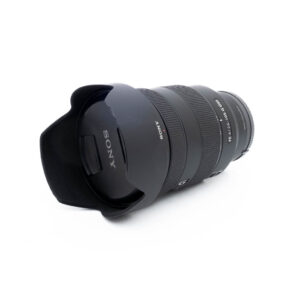 Sony 24-105mm f4 G OSS-3-2 Sony FE 24-105mm f/4 G OSS - Käytetty musta kameraobjektiivi, jossa on vastavalosuoja, näkyy valkoisella taustalla vasemmalle kallistettuna, jotta etuelementti, tuotemerkki ja zoomausmerkinnät tulevat näkyviin.
