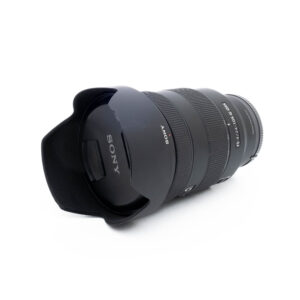 Sony FE 24-105mm f/4 G OSS - Käytetty musta kameraobjektiivi, jossa on vastavalosuoja, näkyy valkoisella taustalla vasemmalle kallistettuna, jotta etuelementti, tuotemerkki ja zoomausmerkinnät tulevat näkyviin.