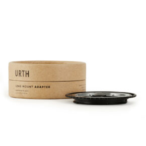 1677857056_417 Pyöreä laatikko, jossa lukee "Urth Contax/Yashica (C/Y) - Canon (EF / EF S) objektiiviadapteri", on mustan metallisen objektiiviasennussovittimen vieressä valkoisella pohjalla.