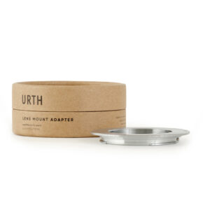 Urth M42 - Canon (EF / EF S) objektiiviadapteri lepää valkoisella pinnalla pyöreän ruskean pahvilaatikon vieressä, jossa lukee URTH LENS MOUNT ADAPTER.