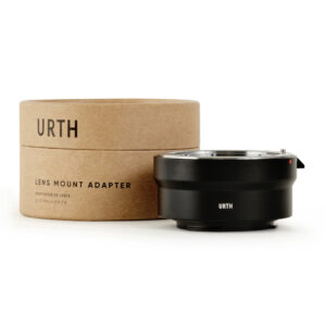 1677857249_063 The cylindrical cardboard box, which reads Urth Pentax K - Canon EF M lens adapter, is next to a black metal lens mount adapter, both against a white background.