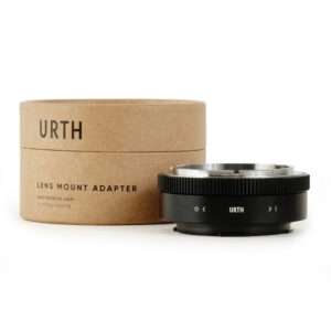 Lieriömäinen pahvilaatikko, jossa lukee "Urth Canon FD - Leica L objektiiviadapteri", on mustan metallisen objektiivisovittimen vieressä, jossa on valkoiset merkinnät ja Urth-logo, molemmat valkoista taustaa vasten.
