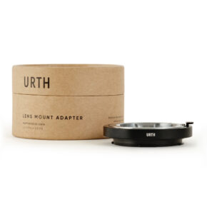 Pahvilaatikko, jossa lukee Urth Leica M - Leica L objektiiviadapteri, on sijoitettu mustan metallisen linssiadapterin viereen, ja molemmat ovat esillä valkoisella pohjalla.