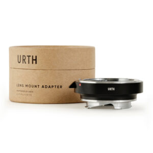 Urth Nikon F - Leica M objektiiviasennussovitin näkyy lieriömäisen pahvipakkauksensa edessä, jossa on merkintä URTH Lens Mount Adapter. Musta sovitin, jossa on hopeisia korostuksia, lepää valkoista taustaa vasten.