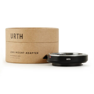 Urth Nikon F - Pentax K objektiiviadapteri näkyy sen lieriömäisen ruskean pahvipakkauksen vieressä, molemmissa on merkintä URTH. Kompaktin mustan sovittimen muotoilussa on metallisia korostuksia.