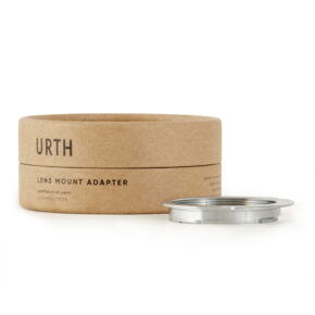 Pyöreä pahvilaatikko, jossa lukee "Urth M42 - Pentax K objektiiviadapteri", näkyy hopeisen metallisen objektiiviasennussovittimen vieressä, molemmat valkoisella pohjalla.