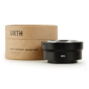 Musta Urth M42 - Sony E objektiiviadapteri istuu sen lieriömäisen ruskean pahvilaatikon vieressä, jossa on merkintä URTH Lens Mount Adapter valkoista taustaa vasten.