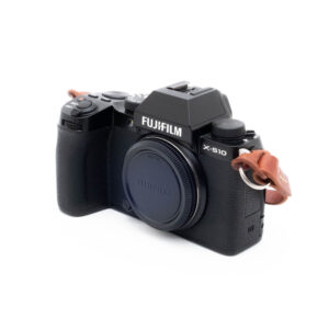 Fujifilm X-S10 (SC 2000, Kunto K5) - Käytetty peilitön kamera, jossa on musta runko, linssinsuojus ja ruskea nahkahihna oikealla puolella valkoista taustaa vasten.