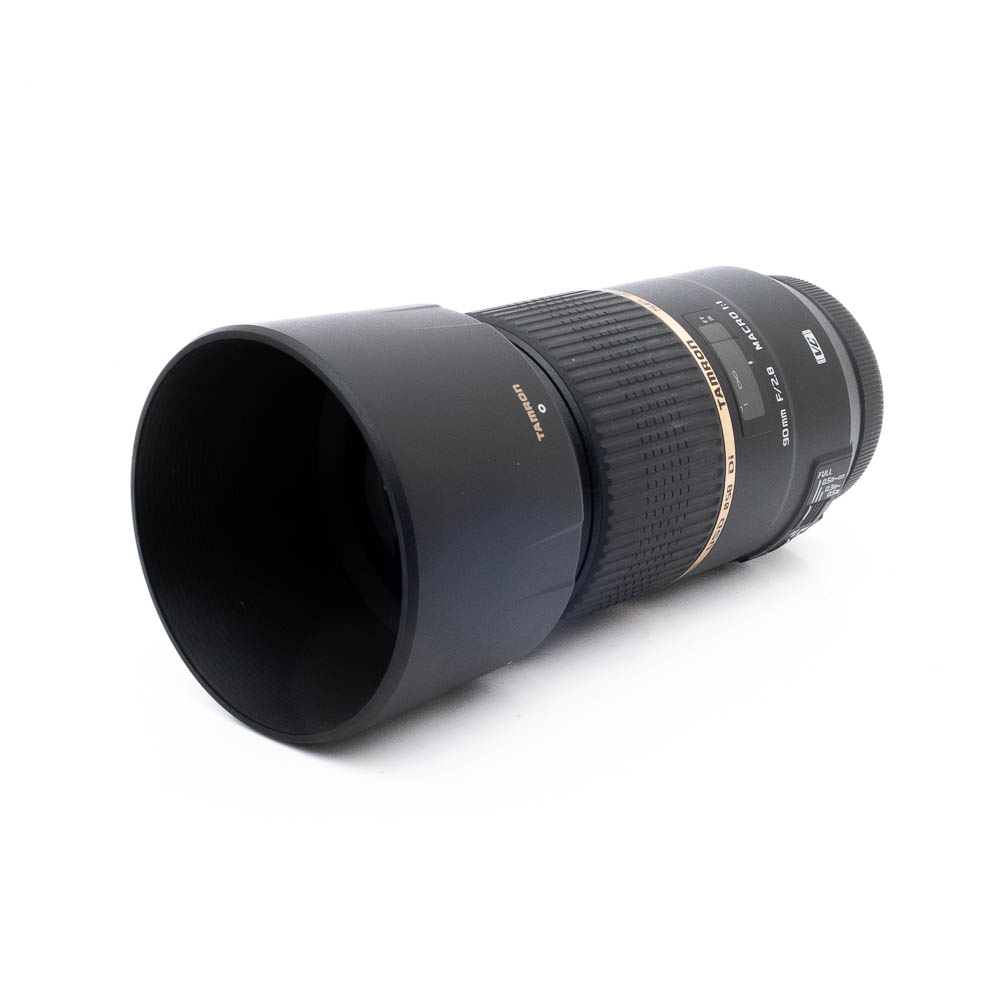 Tamron 90mm f28 Canon-3 Tamron SP 90mm f/2.8 USD Di VC Macro Canon-objektiivi (käytetty, Kunto K5), musta, kultarengas ja vastavalosuoja, kuvattuna kulmassa valkoisella taustalla.