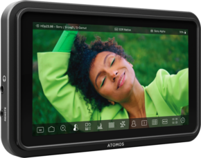Atomos Shinobi II 5,2" -näytön lähikuva, jossa näkyy elävä kuva hymyilevästä henkilöstä, jolla on kiharat hiukset ja vihreä asu ja joka poseeraa toinen käsi kohotettuna pehmeästi sumeaa vihreää taustaa vasten.