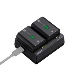 Sony BC-ZD1 Laturi -mustassa akkulaturissa on kaksi kameran akkua, joissa molemmissa on vihreät valot, ja se saa virtansa valkoisesta USB-kaapelista.