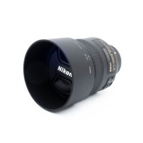 Nikon 85mm f18G-3-2 Nikon AF-S Nikkor 85mm f/1.8 G (Kunto K5) - Käytetty objektiivi, jossa on vastavalosuoja ja suojus, valkoisella pohjalla, jossa on selkeät objektiivin merkinnät ja yksityiskohdat näkyvissä sivuilla.