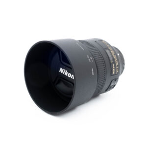 Nikon 85mm f18G-3-2 Nikon AF-S Nikkor 85mm f/1.8 G (Kunto K5) - Käytetty objektiivi, jossa on vastavalosuoja ja suojus, valkoisella pohjalla, jossa on selkeät objektiivin merkinnät ja yksityiskohdat näkyvissä sivuilla.