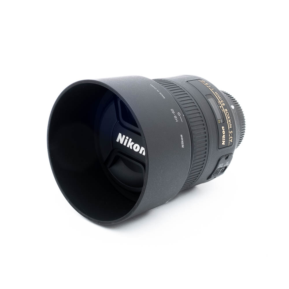 Nikon 85mm f18G-3-2 Nikon AF-S Nikkor 85mm f/1.8 G (Kunto K5) - Käytetty objektiivi, jossa on vastavalosuoja ja suojus, valkoisella pohjalla, jossa on selkeät objektiivin merkinnät ja yksityiskohdat näkyvissä sivuilla.