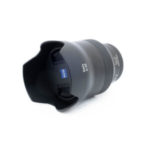 Zeiss Batis 25mm f2 Distagon-3 Mustan värinen Zeiss Batis 25mm f/2 Distagon Sony - Käytetty -objektiivi, jossa on terälehden muotoinen vastavalosuoja ja näkyvä sininen Zeiss-logo tynnyrissä, on kuvassa kyljellään valkoista taustaa vasten.