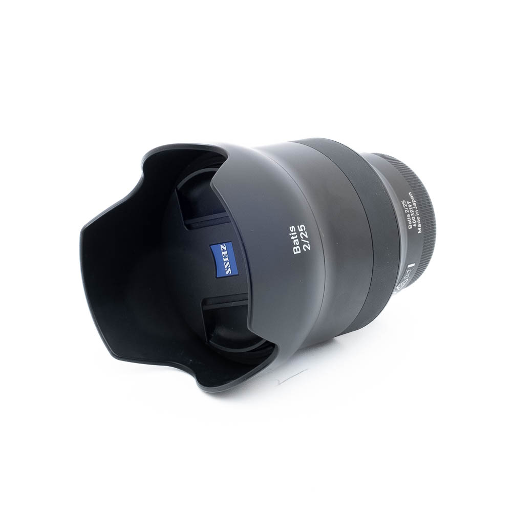 Zeiss Batis 25mm f2 Distagon-3 Mustan värinen Zeiss Batis 25mm f/2 Distagon Sony - Käytetty -objektiivi, jossa on terälehden muotoinen vastavalosuoja ja näkyvä sininen Zeiss-logo tynnyrissä, on kuvassa kyljellään valkoista taustaa vasten.
