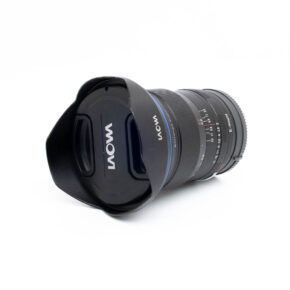 Laowa 15mm f/2 Zero-D Sony - Käytetty -objektiivi, jossa on terälehden muotoinen huppu ja objektiivin suojus, makaa kyljellään valkoista taustaa vasten. Objektiivissa on selvästi merkityt tarkennus- ja aukkoasteikot.