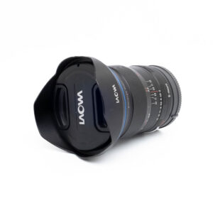 Laowa 15mm f2-3 Laowa 15mm f/2 Zero-D Sony - Käytetty -objektiivi, jossa on terälehden muotoinen huppu ja objektiivin suojus, makaa kyljellään valkoista taustaa vasten. Objektiivissa on selvästi merkityt tarkennus- ja aukkoasteikot.
