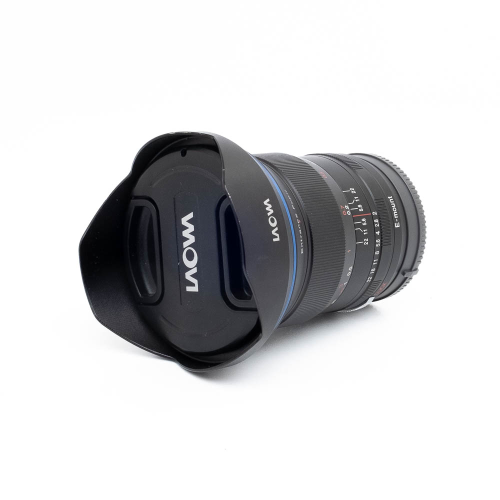 Laowa 15mm f2-3 Laowa 15mm f/2 Zero-D Sony - Käytetty -objektiivi, jossa on terälehden muotoinen huppu ja objektiivin suojus, makaa kyljellään valkoista taustaa vasten. Objektiivissa on selvästi merkityt tarkennus- ja aukkoasteikot.