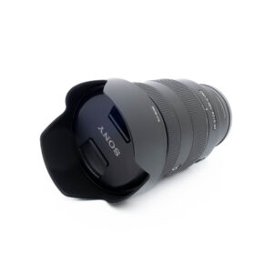 Sony 24-105mm f4 G OSS-3-5 Sony FE 24-105mm f/4 G OSS -objektiivi (käytetty), musta, vastavalosuoja kiinnitettynä, kuvakulmassa valkoisella taustalla. Objektiivin suojuksessa näkyy Sonyn logo.