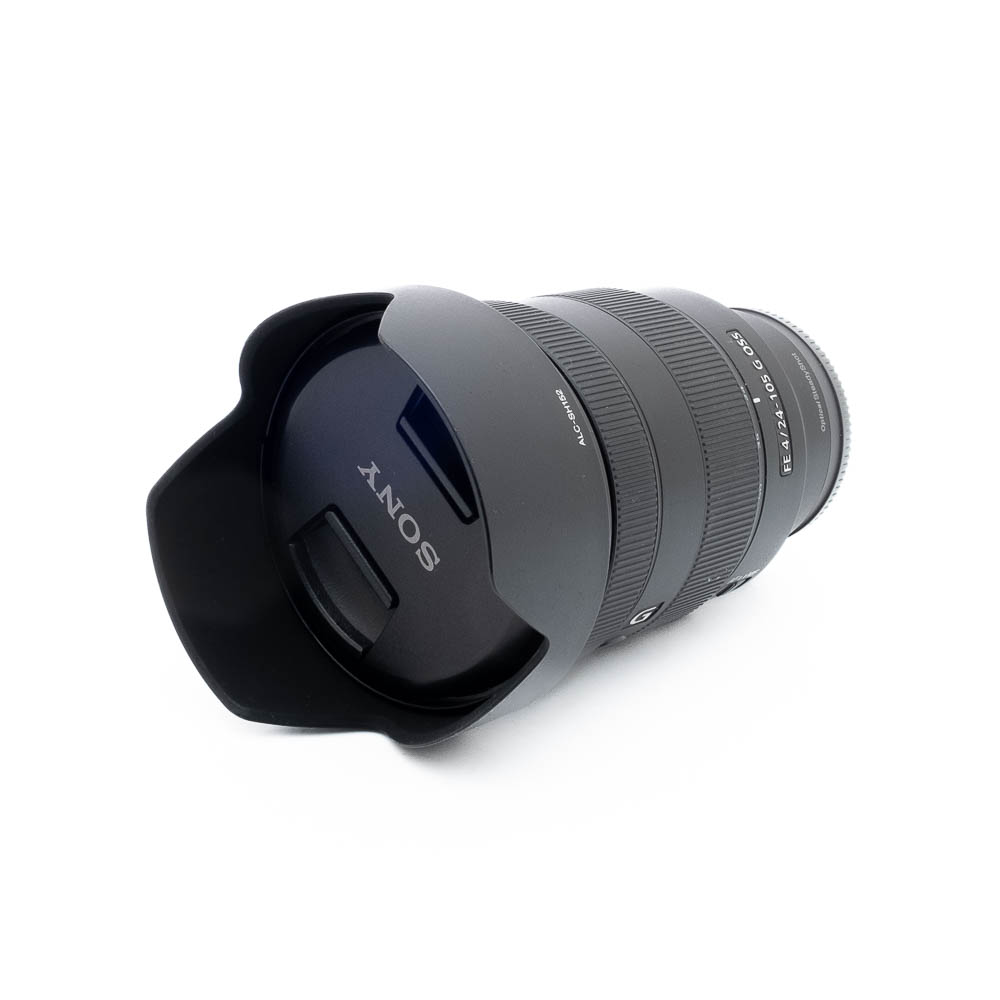 Sony 24-105mm f4 G OSS-3-5 Sony FE 24-105mm f/4 G OSS -objektiivi (käytetty), musta, vastavalosuoja kiinnitettynä, kuvakulmassa valkoisella taustalla. Objektiivin suojuksessa näkyy Sonyn logo.