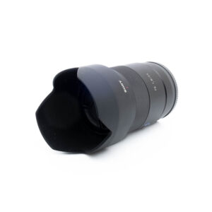 Sony 55mm f18 ZA-3-3 Sony FE Sonnar T* 55mm f/1.8 ZA - Käytetty musta objektiivi, jossa on terälehden muotoinen huppu, kuvattuna kulmasta valkoista taustaa vasten.