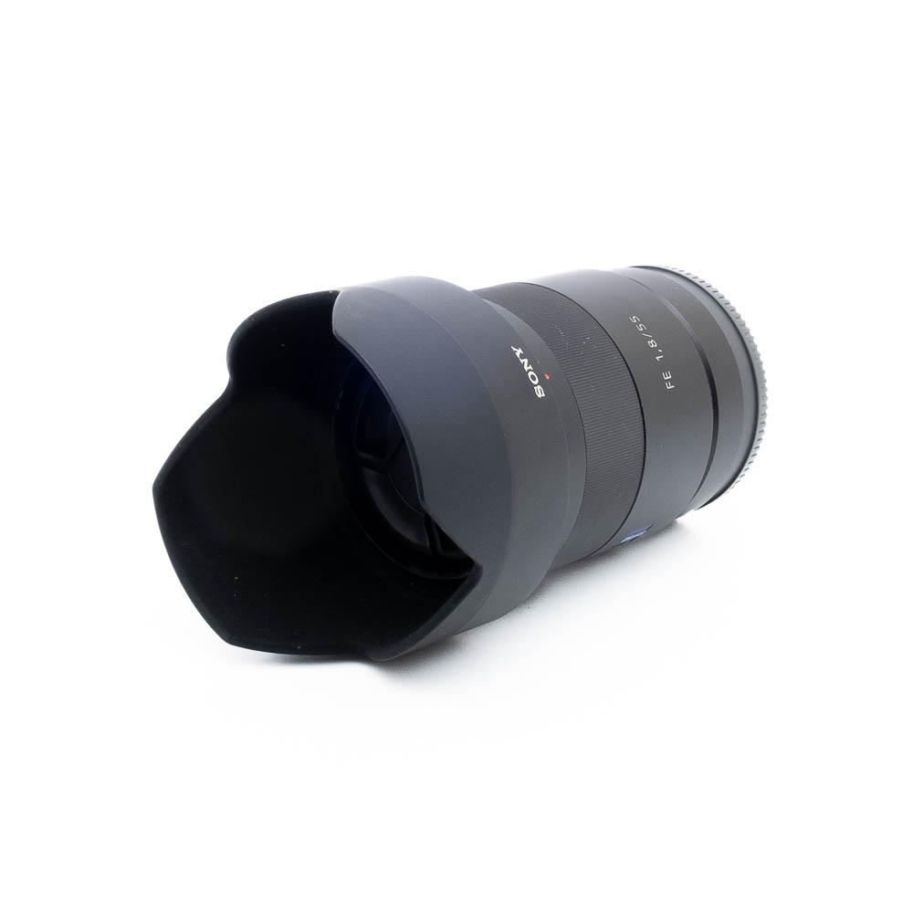 Sony 55mm f18 ZA-3-3 Sony FE Sonnar T* 55mm f/1.8 ZA - Käytetty musta objektiivi, jossa on terälehden muotoinen huppu, kuvattuna kulmasta valkoista taustaa vasten.