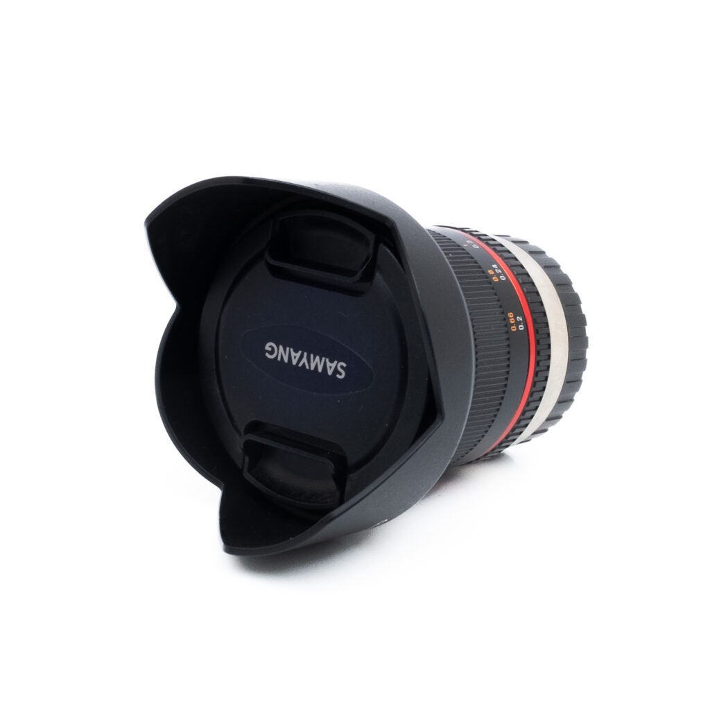 Samy12mm Samyang 12mm f/2 NCS CS Fujifilm (käytetty) kameran objektiivi, jossa on vastavalosuoja ja suojus, makaa kyljellään valkoista taustaa vasten.