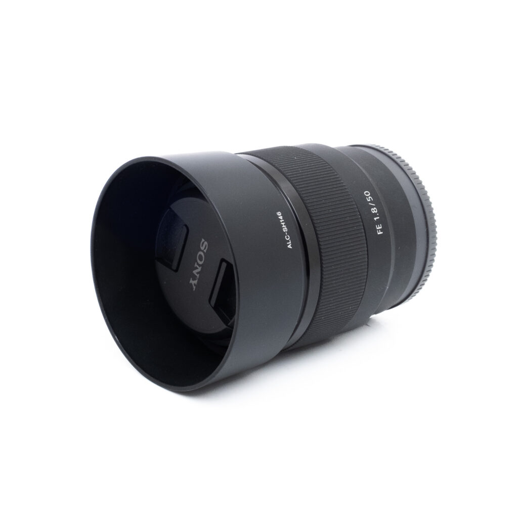 Sony 50mm f18-3 Musta Sony FE 50mm f/1.8 -objektiivi (käytetty), jossa on vastavalosuoja, makaa kyljellään valkoista taustaa vasten.