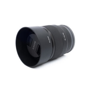 Sony 50mm f18-3 Musta Sony FE 50mm f/1.8 -objektiivi (käytetty), jossa on vastavalosuoja, makaa kyljellään valkoista taustaa vasten.