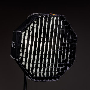 1623067157_424 MagMod Magbox Pro 24 Octa Grid, musta kahdeksankulmainen softbox, jossa on ritilädiffuusori, on asennettu jalustalle tummaa taustaa vasten ammattimaiseen studiovalaistukseen.