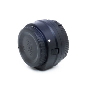 Musta Nikon FTZ II -adapteri (Kunto K5) - Käytetty näkyy kulmassa valkoista taustaa vasten, ja siinä on objektiivin suojus, kuvioidut pintatiedot ja kiinnityspiste.