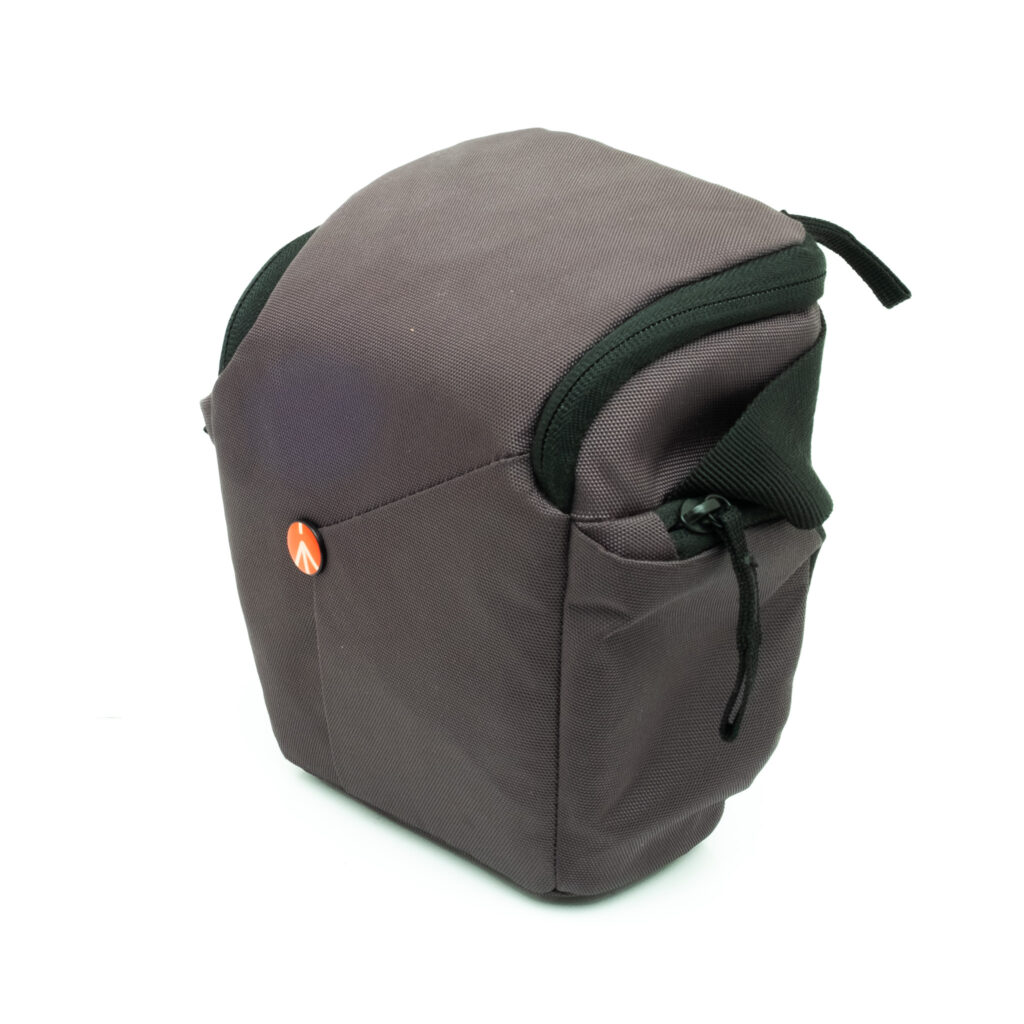 frotto-2 Manfrotto NX Camera Holster - Käytetty on harmaa kameralaukku mustilla korostuksilla ja pienellä oranssilla logolla, jossa on vetoketjullinen yläosa ja sivutaskut. Kuvattu valkoisella pohjalla.