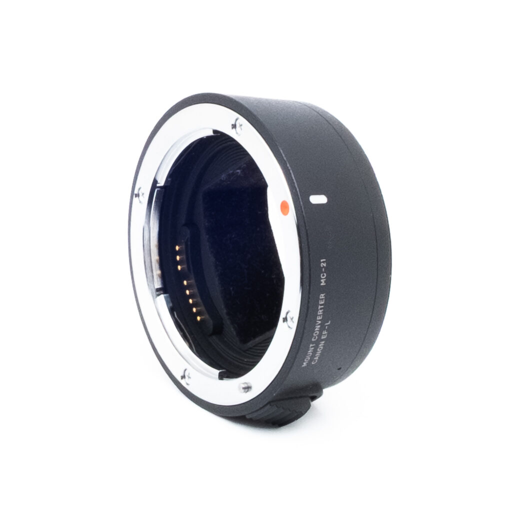 Sigma MC-21-2 Sigma Mount Converter MC-21 (sis. ALV 25,5%) - Käytetty on musta objektiivisovitin, jossa on metalliset kiinnitysrenkaat ja elektroniset koskettimet, jotka näkyvät vinossa valkoisella pohjalla.
