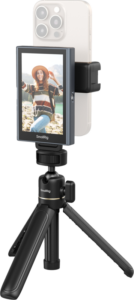 Hattupäinen nainen hymyilee ja poseeraa ulkona, kun hänen älypuhelimensa, joka on kiinnitetty SmallRig 4851 Wireless Video Monitor for Phone (Vlog Kit) -laitteeseen, näyttää hänen kuvansa näytöllä.
