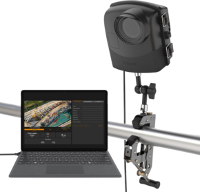 Mustan värinen Brinno BCC2000Plus Construction Camera Bundle on asennettu metallitolppaan ja liitetty kannettavaan tietokoneeseen, jossa näkyy editointiohjelmisto ja ilmakuva venesatamasta.
