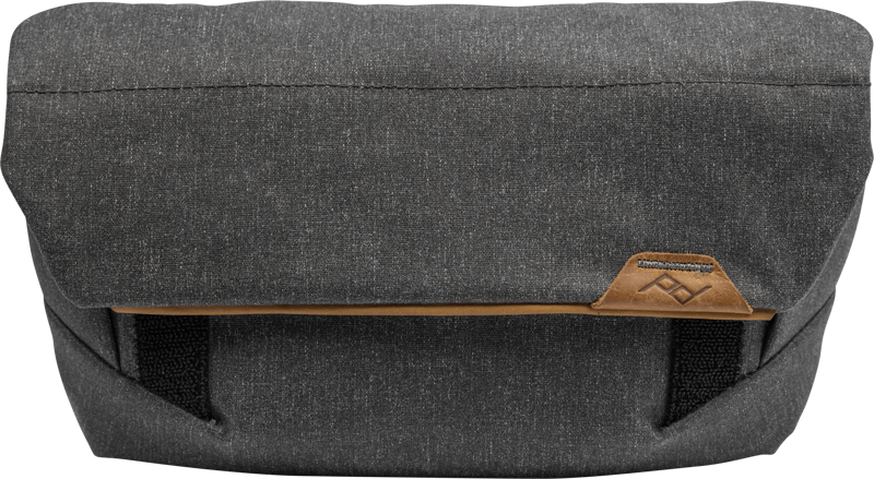 800q-01_123331 Peak Design The Field Pouch v2 in Charcoal on musta kankainen messenger-tyylinen laukku, jossa on taittuva läppä, ruskea nahka-aksentti ja pieni ommeltu logolaastari edessä.