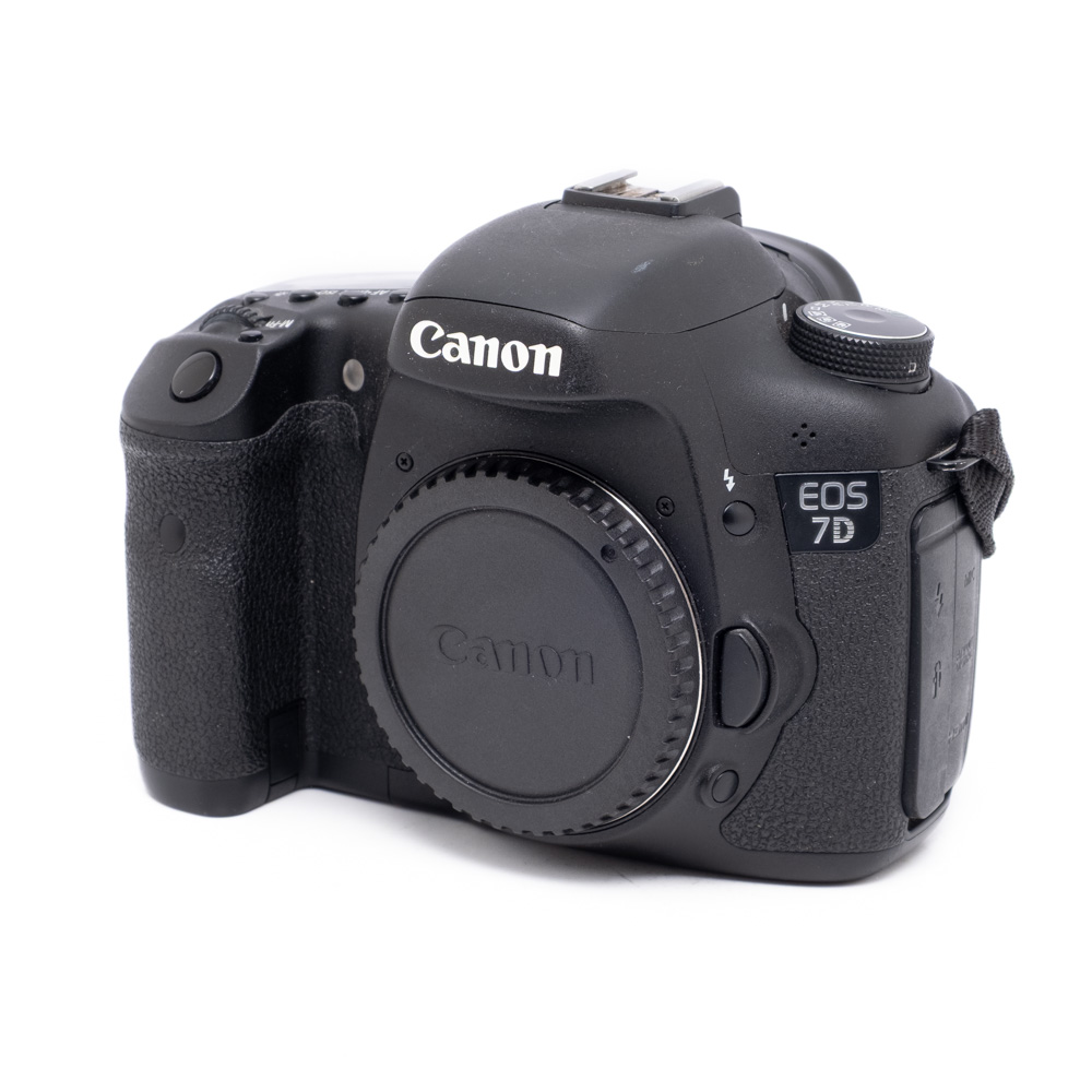 Canon EOS 7D-1 Musta Canon EOS 7D (SC 11000) - Käytetty kamerarunko ilman linssiä valkoisella pohjalla, jossa näkyy etupuoli ja vasen puoli, jossa näkyy kahva ja logo.