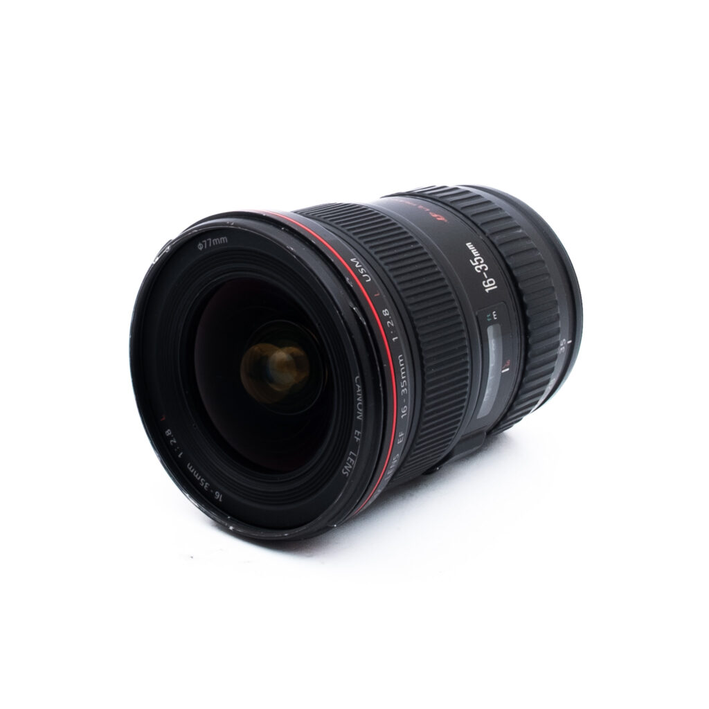 Canon 16-35mm f28 L USM-2 Musta kameran objektiivi, jossa on punavalkoiset merkinnät 16-35mm, lepää valkoisella pohjalla, vinossa kuvattuna niin, että etulasin elementti ja objektiivin rungon yksityiskohdat näkyvät.