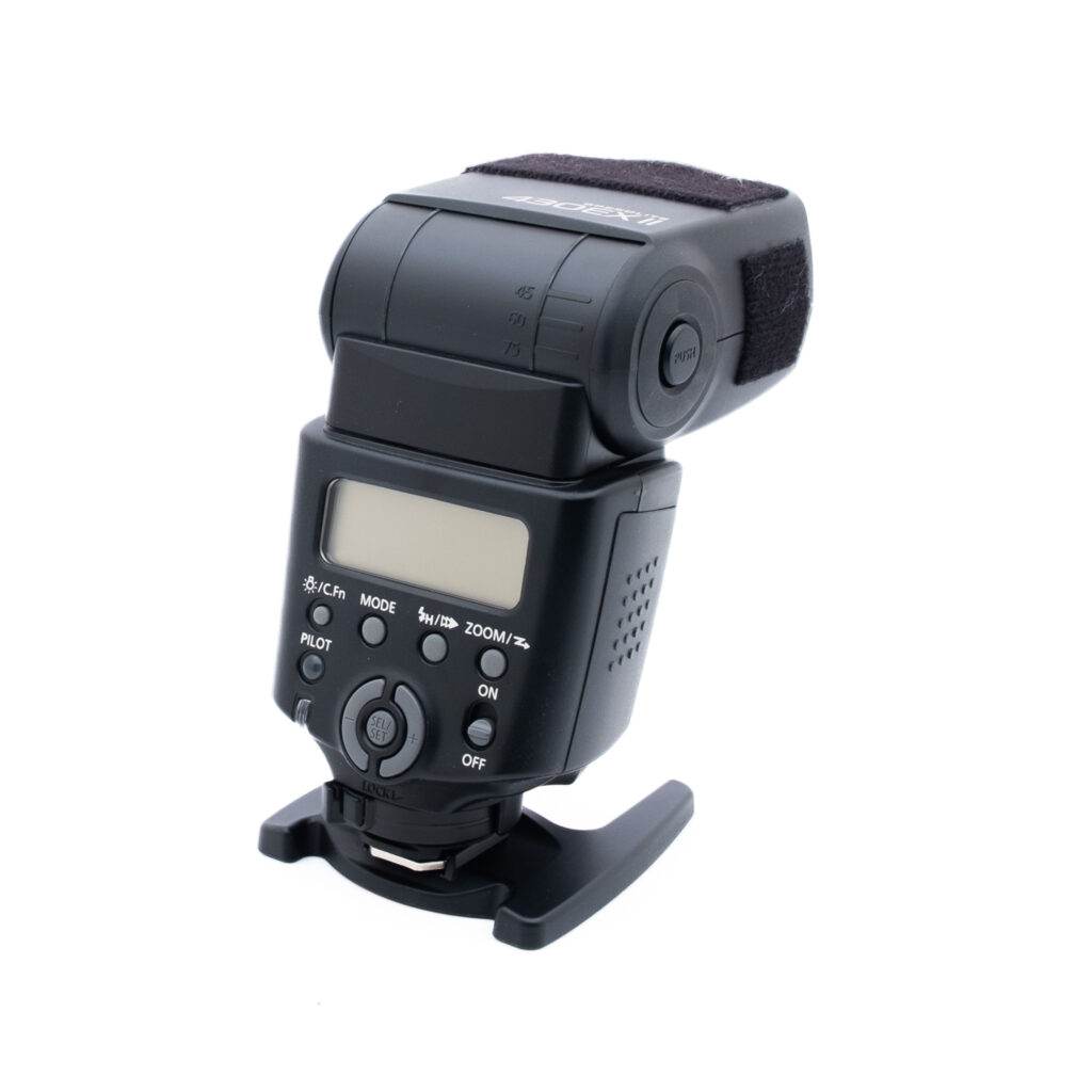 Canon 430EX II-1 Musta kameran ulkoinen salamalaite, jonka takaosassa on digitaaliset säätimet ja painikkeet, seisoo pystyssä valkoisella pohjalla.